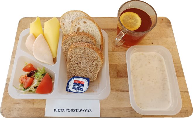 Zestaw śniadaniowy dla diety podstawowej: kilka kromek pieczywa jasnego i ciemnego, plastry żółtego sera i wędliny drobiowej, mały miks sałaty z pomidorem, porcja masła w pojemniku, szklanka herbaty z plastrem cytryny oraz miseczka owsianki na mleku. Pod tacą widoczna etykieta z nazwą diety