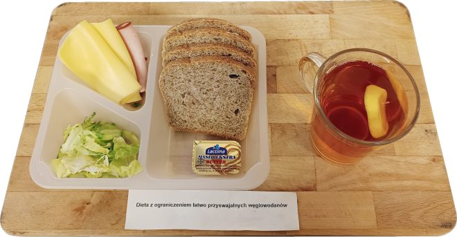 Tacka z posiłkiem szpitalnym w diecie z ograniczeniem łatwo przyswajalnych węglowodanów: kilka kromek pieczywa razowego, plasterki sera żółtego i wędliny, porcja posiekanej sałaty, masło oraz kubek herbaty z plasterkiem cytryny. Na desce leży kartka z napisem „Dieta z ograniczeniem łatwo przyswajalnych węglowodanów”