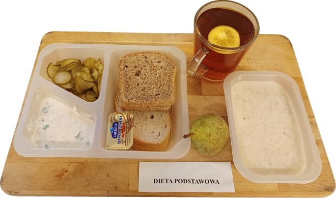 Zestaw śniadaniowy w plastikowej tacce podpisany „Dieta podstawowa”, ustawiony na drewnianej desce. W skład posiłku wchodzą pieczywo pszenne i razowe, masło, twarożek ze szczypiorkiem, plasterki ogórka kiszonego, gruszka, szklanka herbaty z cytryną oraz miseczka białej owsianki