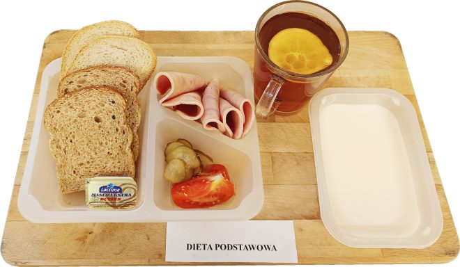 Na tacy znajduje się porcjowane pieczywo, masło, kilka plasterków szynki oraz niewielka porcja ogórków i pomidora. Obok stoi szklanka herbaty z cytryną oraz pojemniczek z jogurtem lub mlecznym deserem. Przed tacą leży etykieta „Dieta podstawowa”