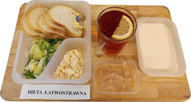 Na drewnianej tacy znajduje się zestaw śniadaniowy w jasnych plastikowych pojemnikach. W największym z nich leżą kromki jasnego pieczywa oraz mała porcja masła. Obok znajduje się pojemnik z mieszanką sałat. W kolejnym pojemniku umieszczono jajecznicę. Przed nim stoi niewielkie pudełko z musem jabłkowym. Po prawej stronie widoczny jest pojemnik z płynną kaszą manną lub mleczną zupą. W tle stoi szklanka herbaty z plasterkiem cytryny. Przed zestawem umieszczono etykietę „Dieta łatwostrawna”