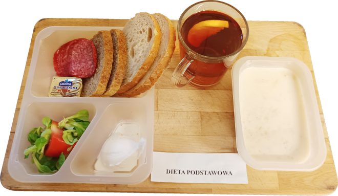 Śniadanie w diecie podstawowej: tacka z pieczywem, plasterkami salami, masłem, sałatką z pomidorem i zieloną sałatą oraz porcją białego twarożku. Obok stoi kubek herbaty z cytryną i pojemnik z białą zupą mleczną. Na tacy widoczny podpis «Dieta podstawowa