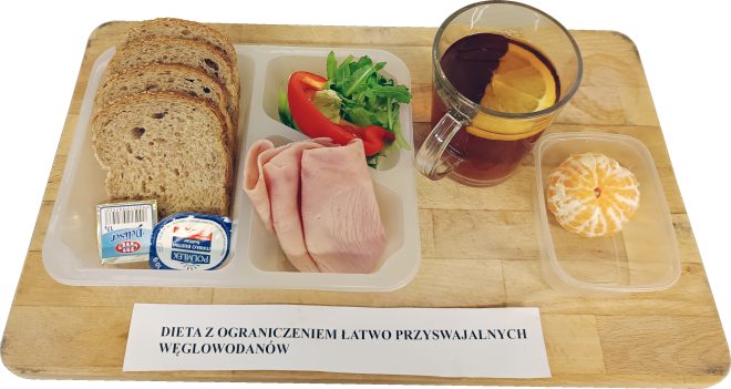 Taca z zestawem śniadaniowym w diecie z ograniczeniem łatwo przyswajalnych węglowodanów: kilka kromek pieczywa pełnoziarnistego, masło w porcji jednostkowej, serek biały homogenizowany, plastry chudej wędliny oraz porcja warzyw – sałata, rukola i pasek czerwonej papryki. Obok znajduje się szklanka herbaty z cytrusowym plasterkiem i osobny pojemniczek z obranym mandarynkowym owocem