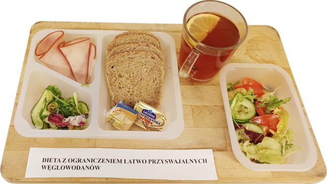 Zestaw śniadaniowy na tacy: plasterki chudej wędliny, porcja zielonej sałatki, kilka kromek pełnoziarnistego pieczywa, dwie małe porcje masła i serka, oddzielna miseczka z sałatką z pomidora i ogórka oraz szklanka herbaty z cytryną. Przed tacą leży karteczka z napisem „Dieta z ograniczeniem łatwo przyswajalnych węglowodanów”