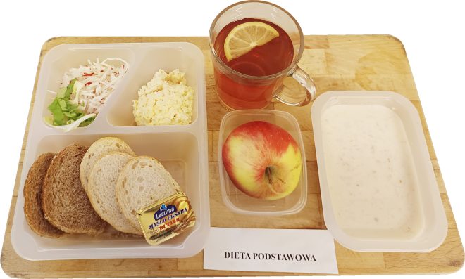 Śniadanie w diecie podstawowej podane na plastikowej tacy. W zestawie znajdują się kanapki z pieczywem jasnym i razowym oraz porcją masła, pasta jajeczna, surówka z białej rzodkwi z dodatkiem sałaty, a także jabłko. Obok tacy stoi szklanka herbaty z plasterkiem cytryny oraz miseczka z mleczną zupą. Przed tacą widoczna jest kartka z napisem „Dieta podstawowa”