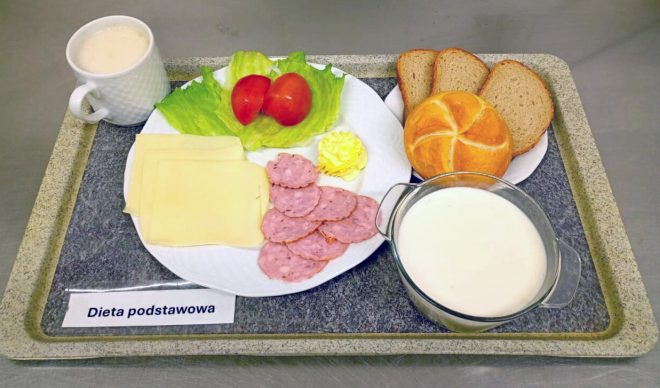Na tacy znajduje się śniadanie w diecie podstawowej. Na talerzu ułożono plasterki żółtego sera, kawałki wędliny, liść sałaty, pomidora i porcję masła. Obok leży kajzerka i kilka kromek chleba razowego. W szklanym naczyniu znajduje się mleko, a w filiżance napój mleczny lub kawa z mlekiem. Na tacy widoczna jest również karteczka z napisem „Dieta podstawowa”
