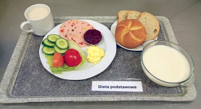 Zestaw śniadaniowy w diecie podstawowej. Na tacy znajduje się talerz z plastrami kolorowej galaretowanej wędliny, porcją konfitury, plasterkami ogórka i pomidora, liściem sałaty oraz porcją masła. Obok leżą kromki chleba i bułka kajzerka. Na tacy znajduje się również miseczka z białą zupą mleczną oraz kubek z jasnym napojem