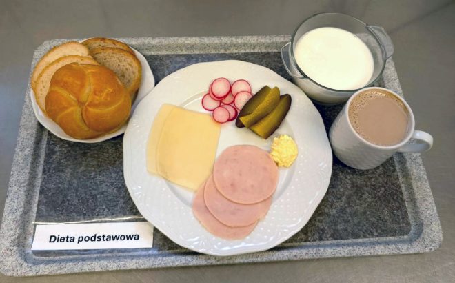 Śniadanie w diecie podstawowej. Na tacy znajduje się talerz z trzema plastrami szynki, dwoma plastrami sera żółtego, plastrami rzodkiewki, kawałkami ogórka kiszonego i porcją masła. Obok ułożono bułkę i kromki chleba. Na tacy stoi również miseczka z mlekiem oraz kubek z napojem kakaowym
