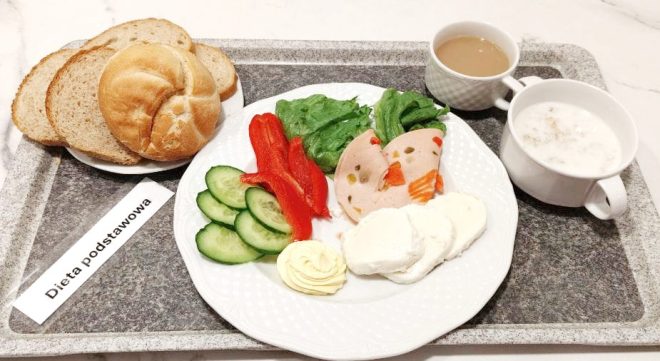 Na tacy z napisem „Dieta podstawowa” znajduje się śniadanie. Na talerzu podano plasterki ogórka, paski czerwonej papryki, liście sałaty, plasterki drobiowej wędliny z warzywami, kawałki białego sera oraz porcję masła. Obok leżą trzy kromki pieczywa i bułka kajzerka. W filiżance znajduje się kawa z mlekiem, a w miseczce — płatki owsiane na mleku