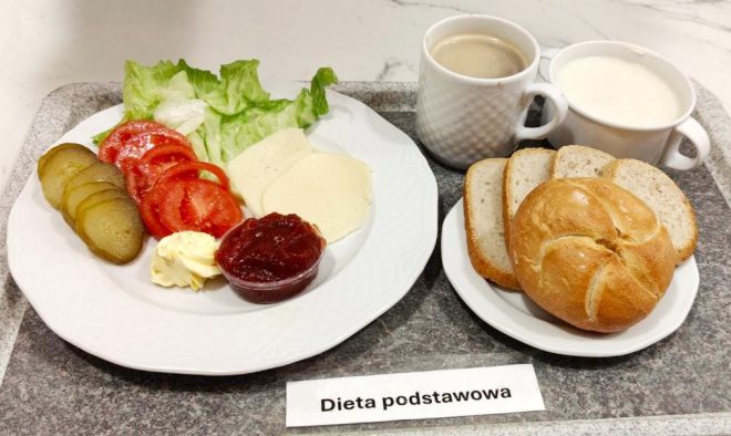 Na tacy śniadanie złożone z pieczywa pszennego i razowego, sałaty, plasterków pomidora, ogórka kiszonego, sera żółtego, masła oraz dżemu. Do zestawu filiżanka kawy z mlekiem i kubek mleka. Na tacy widoczna karteczka z napisem „Dieta podstawowa”