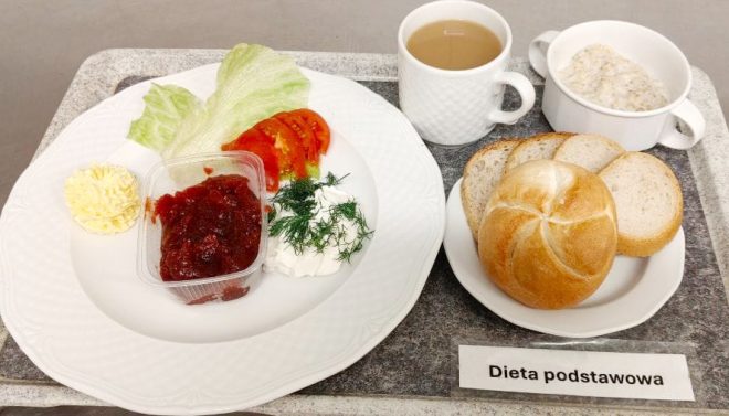 Na tacy zestaw śniadaniowy: pieczywo jasne i bułka kajzerka, płatki owsiane w miseczce, kawa z mlekiem, masło, biały ser z koperkiem, sałata, pomidor, oraz porcja dżemu. Na tacy etykieta „Dieta podstawowa”