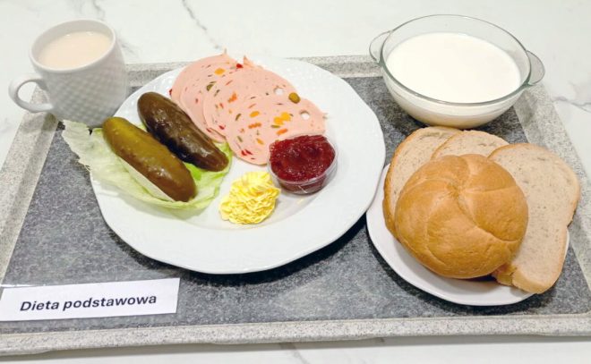 Śniadanie w diecie podstawowej podane na tacy: plasterki wędliny drobiowej z warzywami, dwa ogórki kiszone na liściu sałaty, porcja masła oraz niewielka porcja dżemu. Obok znajdują się kromki pieczywa jasnego i bułka pszenna, miseczka z mlekiem oraz kubek z kawą z mlekiem