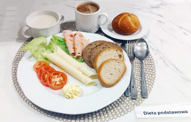 Talerz ze śniadaniem w diecie podstawowej: plastry pomidora, liść sałaty, dwa plastry sera, dwa plastry wędliny drobiowej oraz kromki pieczywa jasnego i razowego. Obok na tacy stoją kubek z kawą z mlekiem, miseczka z białą kawą zbożową oraz mała bułka pszenna podana na talerzyku