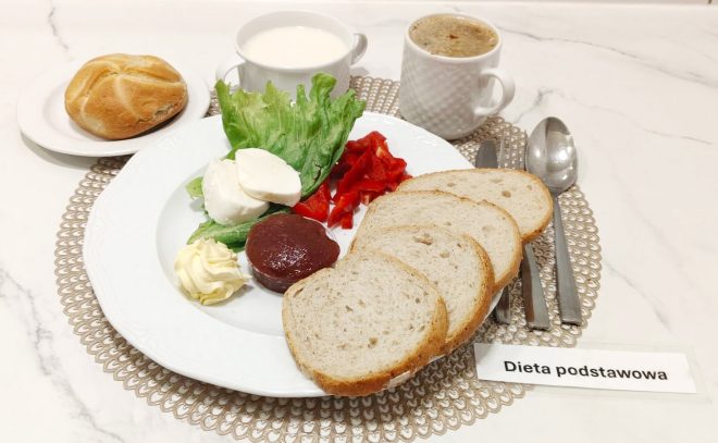 Śniadanie w diecie podstawowej: kilka kromek białego pieczywa, masło, plasterki mozzarelli, galaretka owocowa, sałata i paski czerwonej papryki. Obok bułka kajzerka, kubek kawy z mlekiem i kubek białej kawy