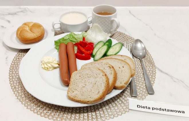 Na dużym talerzu znajdują się parówki, plasterki ogórka i papryki, liść sałaty oraz masło. Dookoła ułożono kilka kromek chleba. Obok stoi kubek z kawą z mlekiem, a z boku bułka na małym talerzyku. Przed zestawem widoczna jest etykieta „Dieta podstawowa”