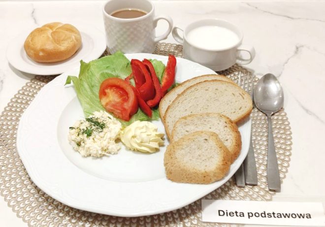 Na talerzu podano świeże warzywa — sałatę, pomidora i paprykę — porcję twarożku ze szczypiorkiem oraz masło. Obok znajdują się kromki pieczywa, filiżanka kawy z mlekiem, miseczka z białym napojem oraz bułka podana na osobnym talerzyku. Widoczna kartka z napisem „Dieta podstawowa”