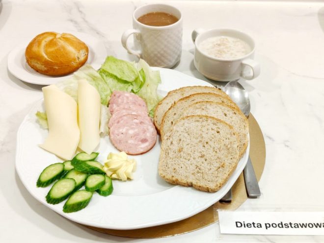 Na talerzu ułożono plasterki sera i wędliny, sałatę, masło oraz pokrojonego ogórka. Obok znajdują się kromki pieczywa, filiżanka kawy z mlekiem, miseczka z mlekiem lub zupą mleczną oraz bułka. Z prawej strony widnieje oznaczenie „Dieta podstawowa”