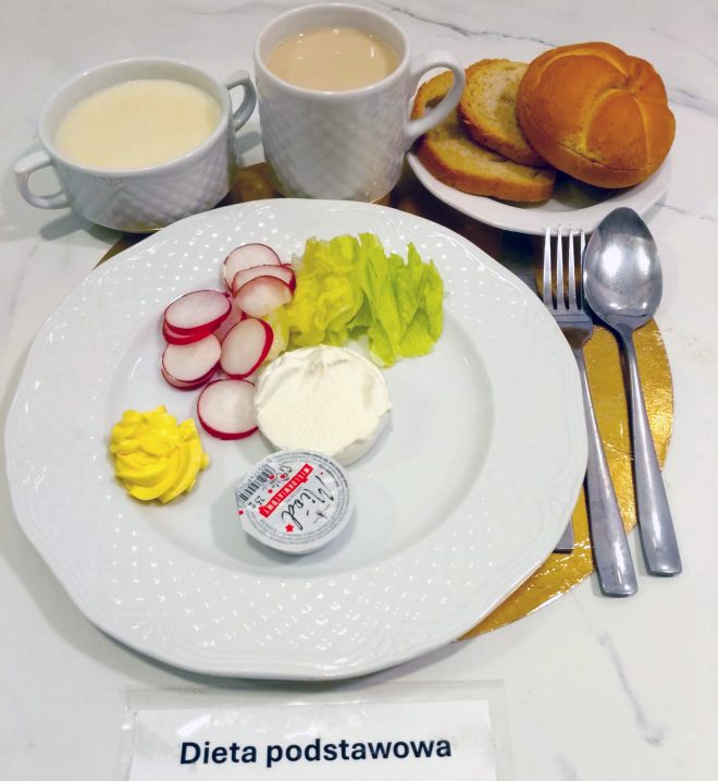 Na talerzu znajduje się porcja twarogu, masło, plastry rzodkiewki oraz liście sałaty. Obok ustawiono filiżankę kawy z mlekiem, miseczkę z zupą mleczną oraz talerzyk z bułką i kromkami pieczywa. Całość podano z etykietą „Dieta podstawowa”