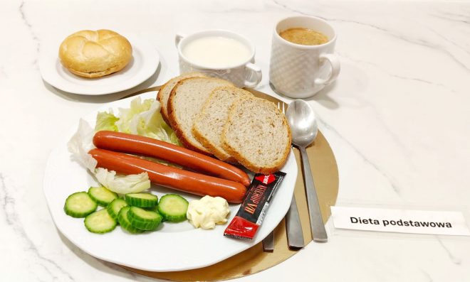 Śniadanie na diecie podstawowej: dwie parówki z dodatkami — ogórek, sałata, masło i ketchup, podane z kromkami chleba oraz bułką; do picia mleko i kawa