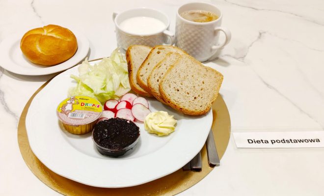 Śniadanie na talerzu: trzy kromki pieczywa, liście sałaty, plasterki rzodkiewki, porcja masła, porcja dżemu oraz serek typu „DrobiPik”. Obok znajduje się bułka na osobnym talerzyku, kubek kawy z mlekiem oraz miseczka białego jogurtu. Po prawej stronie widoczna karteczka z napisem „Dieta podstawowa”