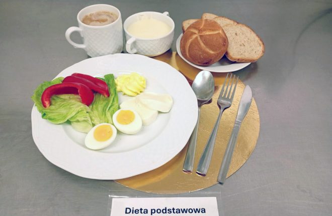 Śniadanie w diecie podstawowej: talerz z liśćmi sałaty, paskami czerwonej papryki, jajkiem gotowanym na twardo, plastrami białego sera i porcją masła. Z tyłu widoczny kubek kawy z mlekiem oraz miseczka z białą zupą mleczną. Zestaw uzupełnia pieczywo — bułka i kromki chleba — podane na małym talerzyku. Obok leżą sztućce. Całość umieszczona na złotej podkładce z etykietą „Dieta podstawowa”