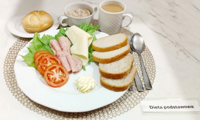 Śniadanie w ramach „Diety podstawowej” podane na białym talerzu. Na talerzu znajdują się liście sałaty, plastry pomidora, zwinięte plasterki wędliny, ser żółty, porcja masła oraz kilka kromek jasnego pieczywa. W tle ustawiono filiżankę kawy z mlekiem, miseczkę owsianki oraz bułkę kajzerkę na osobnym talerzyku