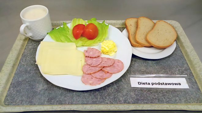 Na zdjęciu śniadanie w ramach diety podstawowej, ułożone na tacy. Na talerzu znajdują się plasterki wędliny, żółty ser, masło, liść sałaty i kawałki pomidora. Obok leżą kromki białego pieczywa oraz kubek z ciepłym napojem mlecznym. Przed zestawem umieszczono karteczkę z napisem „Dieta podstawowa”