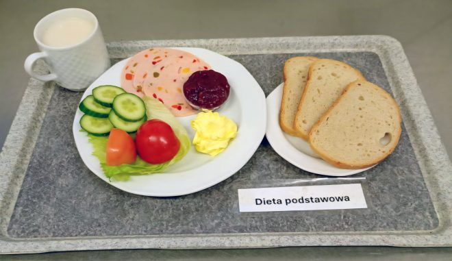 Na tacy z napisem „Dieta podstawowa” znajduje się talerz z kilkoma plastrami wędliny z papryką, porcją masła, galaretką lub konfiturą, oraz warzywami – plasterkami ogórka, liściem sałaty i kawałkami pomidora. Obok leży talerzyk z trzema kromkami jasnego pieczywa i kubek z napojem mlecznym lub kawą z mlekiem
