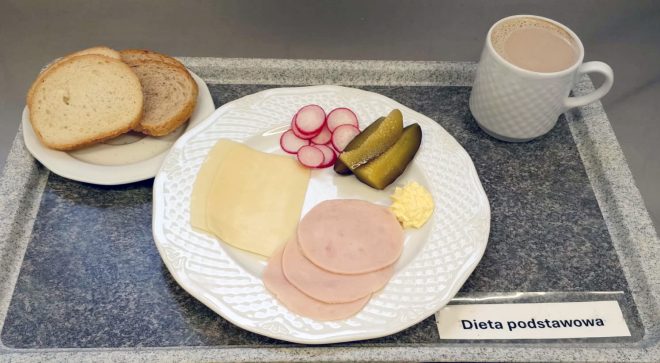 Zestaw śniadaniowy w diecie podstawowej. Na talerzu znajdują się trzy plasterki szynki, dwa plastry sera żółtego, plasterki rzodkiewki, dwa kawałki ogórka kiszonego oraz porcja masła. Obok położono talerzyk z kromkami chleba pszennego i żytniego. Na tacy stoi kubek z kakao