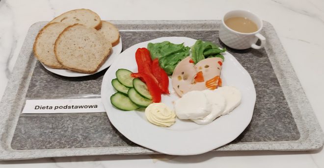 Na tacy z napisem „Dieta podstawowa” znajduje się śniadanie. Na talerzu ułożono kilka plastrów zielonego ogórka, kawałki czerwonej papryki, liście sałaty, plasterki drobiowej wędliny z warzywami, porcję białego sera oraz masło. Obok talerza stoi filiżanka z kawą z mlekiem, a na osobnym talerzyku położono trzy kromki jasnego pieczywa