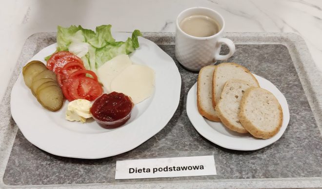 Na tacy z napisem „Dieta podstawowa” znajduje się śniadanie: na białym talerzu leżą plastry pomidora, ogórka kiszonego i sera żółtego, liście sałaty, porcja masła oraz mały pojemniczek z dżemem. Obok stoi kubek z kawą z mlekiem i mały talerzyk z trzema kromkami białego pieczywa. Całość ustawiona jest na szarej tacy