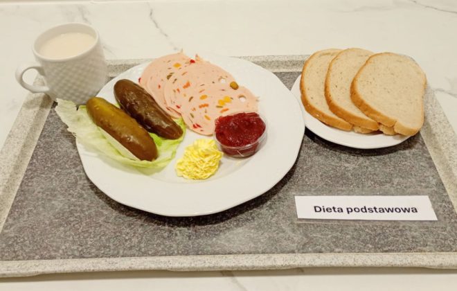 Na tacy śniadanie składające się z trzech kromek białego chleba, plasterków wędliny drobiowej z warzywami, dwóch ogórków kiszonych, porcji masła i dżemu w kubeczku. Obok filiżanka mleka. Na tacy widoczna etykieta „Dieta podstawowa”