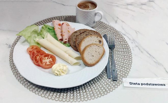 Talerz ze śniadaniem w diecie podstawowej: kilka plastrów pomidora, porcja masła, liść sałaty, dwa plastry sera, dwa plastry wędliny drobiowej oraz kromki pieczywa jasnego i razowego. Obok stoi kubek z kawą. Całość podana na jasnym, dekoracyjnym podkładzie