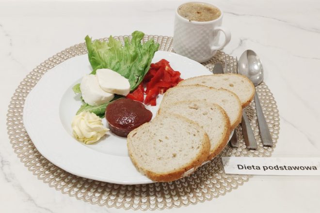Śniadanie w diecie podstawowej to lekki i zbilansowany posiłek, który dostarcza energii na początek dnia. Na talerzu znajdują się świeże warzywa, biały ser, masło, pieczywo oraz słodki akcent w postaci dżemu. Do posiłku serwowana jest kawa zbożowa lub kawa słaba – zgodnie z zaleceniami żywieniowymi