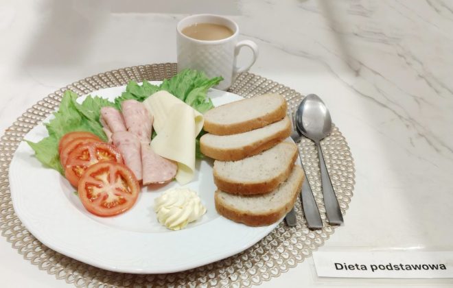 Śniadanie w ramach „Diety podstawowej”, podane na białym talerzu. Na talerzu znajdują się: liście sałaty, plastry pomidora, zwinięte plastry wędliny, ser żółty oraz porcja masła. Obok leży kilka kromek jasnego pieczywa. W tle stoi filiżanka kawy z mlekiem, a z prawej strony ułożone są łyżeczka i łyżka