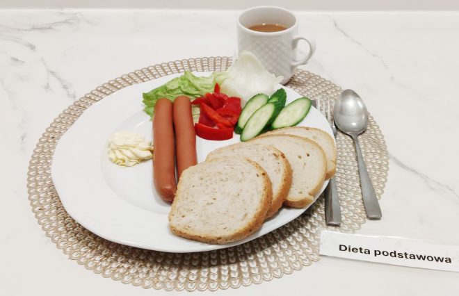 Śniadanie składa się z parówek, plasterków pomidora, ogórka i papryki, porcji masła, liścia sałaty oraz kilku kromek pieczywa. Obok ustawiono kubek z kawą. Zestaw znajduje się na jasnej podkładce z etykietą „Dieta podstawowa”