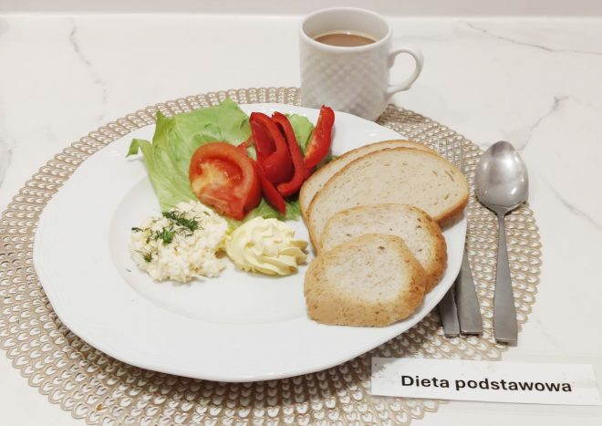 Zestaw śniadaniowy w diecie podstawowej. Na białym talerzu podanym na dekoracyjnej, ażurowej podkładce znajdują się warzywa: liść sałaty, plasterki pomidora oraz paseczki czerwonej papryki. Obok położono porcję twarożku ze szczypiorkiem oraz rozetkę masła. Na talerzu leżą także cztery kromki jasnego pieczywa. Zestaw uzupełnia kubek kawy z mlekiem. Po prawej stronie znajdują się sztućce. Przed talerzem widoczna jest etykieta z napisem ‘Dieta podstawowa’