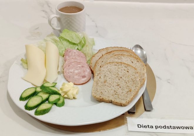 Talerz ze śniadaniem: trzy kromki pieczywa, plasterki ogórka, dwa plastry sera żółtego, kilka plasterków wędliny, liście sałaty oraz porcja masła. Obok kubek z kawą z mlekiem i napis ‘Dieta podstawowa’