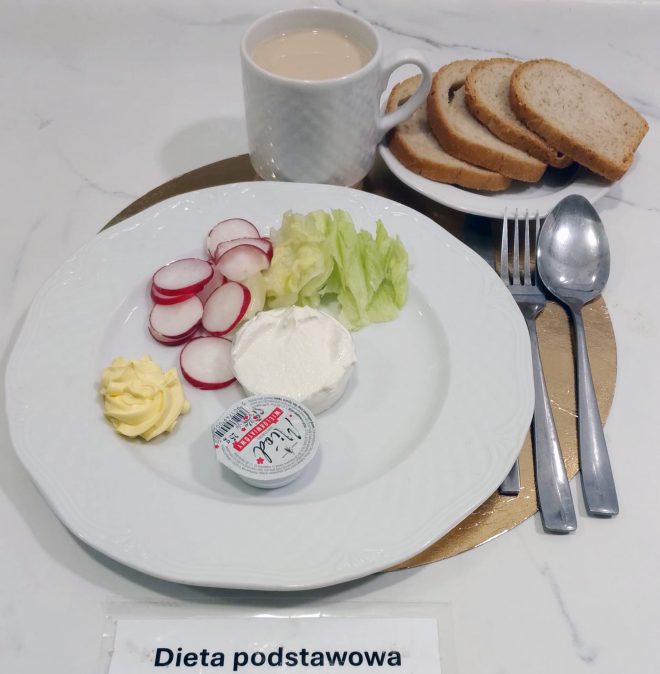 Na białym talerzu ułożono plasterki rzodkiewki, liście sałaty, porcję białego twarogu oraz rozetkę masła. Obok leży mały kubeczek serka. W tle znajduje się filiżanka kawy z mlekiem oraz talerzyk z kilkoma kromkami jasnego pieczywa. Zestaw nakryty jest sztućcami i opisany etykietą „Dieta podstawowa”