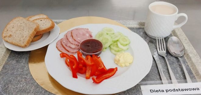 Na tacy podano talerz z kilkoma plastrami wędliny, pokrojonym ogórkiem, paskami czerwonej papryki, porcją masła i małym kubeczkiem konfitury. Obok leży talerzyk z trzema kromkami pieczywa oraz kubek z kawą z mlekiem. Przy tacy umieszczono komplet sztućców i etykietę „Dieta podstawowa”