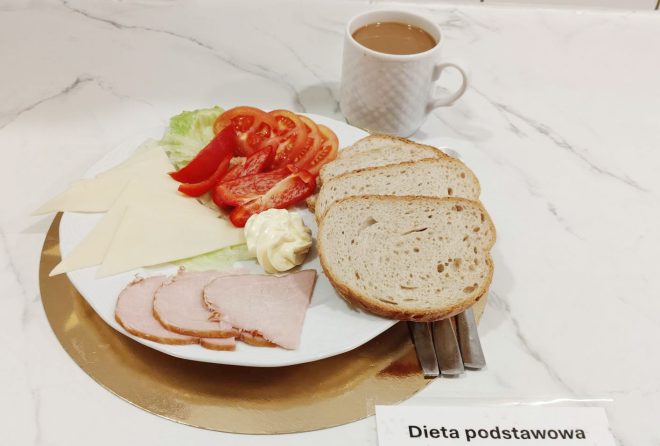 Talerz śniadaniowy z kilkoma plastrami chleba, sałatą, plastrem pomidora i czerwonej papryki, żółtym serem, wędliną oraz porcją majonezu. Obok stoi kubek z kawą z mlekiem. Przed zestawem znajduje się karteczka z napisem „Dieta podstawowa”