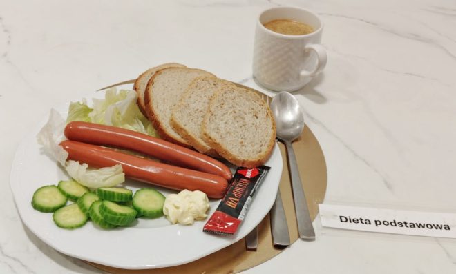 Na białym talerzu umieszczonym na złotej podkładce znajdują się dwie parówki na liściach sałaty, plasterki świeżego ogórka, porcja majonezu oraz trzy kromki pieczywa. Obok leży saszetka ketchupu. W tle stoi biała filiżanka z kawą, a po prawej stronie widoczna jest etykieta z napisem Dieta podstawowa. Przy talerzu ułożono łyżkę i nóż