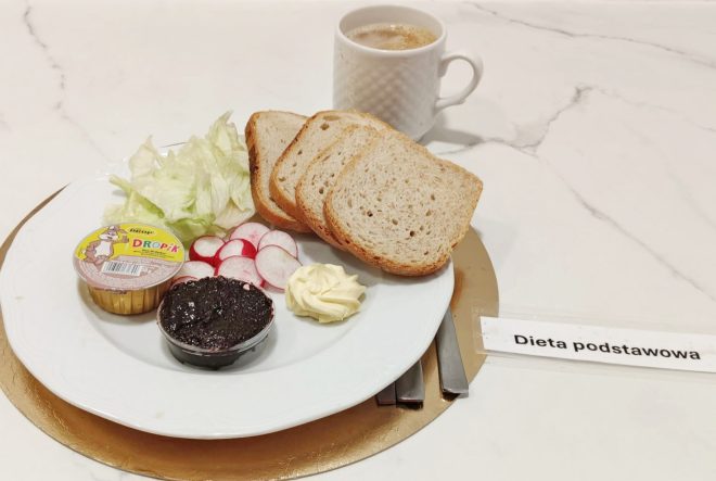 Śniadanie w diecie podstawowej: talerz z kilkoma kromkami jasnego pieczywa, sałatą lodową, plasterkami rzodkiewki, porcją masła oraz dwoma małymi opakowaniami — pasztetu Dropek i czarnej pasty kanapkowej. W tle stoi kubek kawy z mlekiem. Całość ułożona na złotej podkładce z podpisem „Dieta podstawowa”