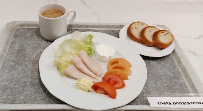 „Śniadanie w diecie podstawowej – pieczywo pszenne z dodatkiem wędliny drobiowej, pomidora, listków sałaty i porcji twarożku. Do zestawu podano kawę z mlekiem.