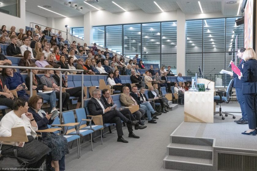 Duża sala audytoryjna wypełniona uczestnikami konferencji. Słuchacze siedzą w kilku rzędach, część robi notatki lub spogląda w stronę sceny. Na podium po prawej stronie dwie osoby prowadzą wystąpienie przy mównicy i ekranie projekcyjnym