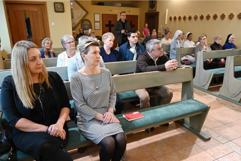 Grupa kobiet i mężczyzn siedzi w ławkach kaplicy podczas nabożeństwa lub spotkania modlitewnego. W pierwszym rzędzie widoczne są dwie kobiety, a za nimi kolejne osoby, w tym siostry zakonne w habitach. Wszyscy słuchają uważnie, panuje skupiona i spokojna atmosfera