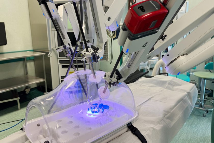 Ramiona robota da Vinci pracujące na trenażerze laparoskopowym. Przez przezroczystą kopułę widać pole treningowe oświetlone fioletowym światłem LED. Robot wykonuje precyzyjne ruchy narzędziami, symulując procedurę chirurgiczną