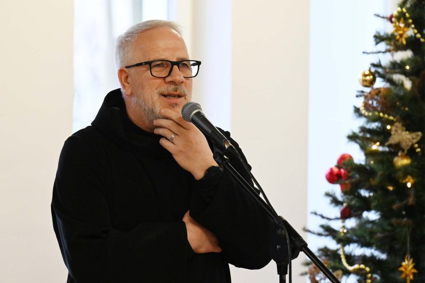 Duchowny w czarnym habicie przemawia do mikrofonu; ma okulary i siwą brodę, jedną rękę opiera pod brodą, a w tle widać udekorowaną choinkę z bombkami i światełkami