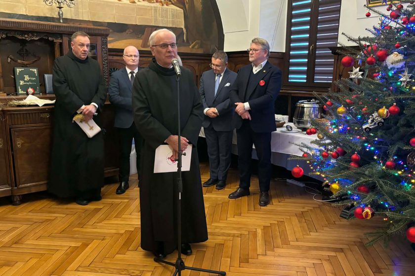Duchowny przemawiający przy mikrofonie podczas spotkania opłatkowego w zabytkowej sali, w tle zaproszeni goście oraz udekorowana choinka