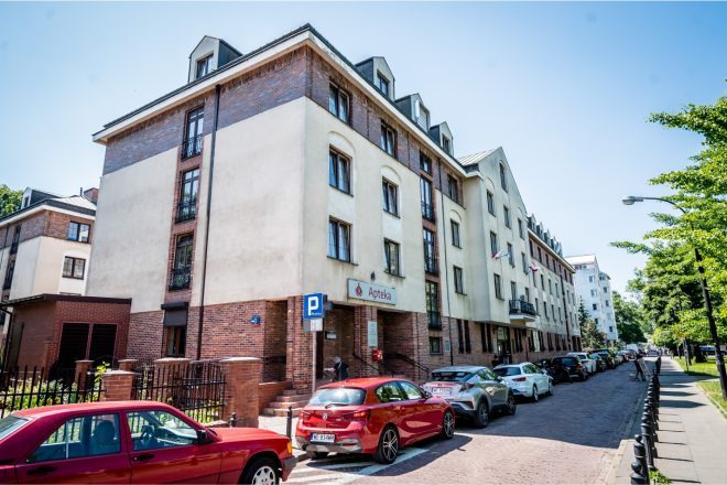Budynek Przychodni z apteką na parterze, widziany od strony ulicy. Przed wejściem zaparkowane samochody, wzdłuż ulicy chodnik i zieleń
